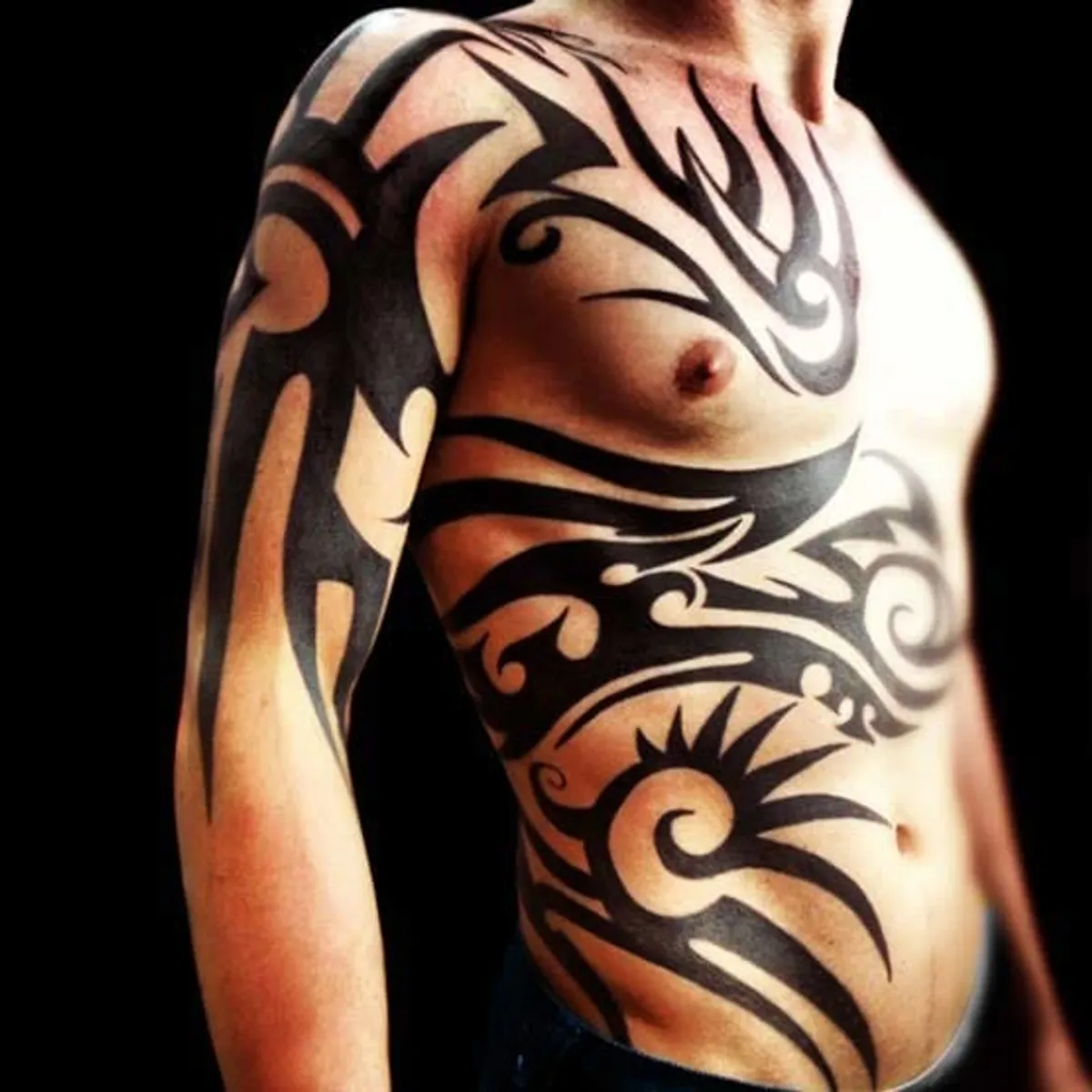 A close-up of a man's upper body displaying intricate black tribal tattoos on his arm and torso against a dark background.